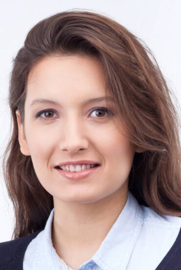 A woman with long brown hair and a bright smile is looking directly at the camera. She is wearing a light blue collared shirt under a dark sweater. The background is plain white, emphasizing her features.