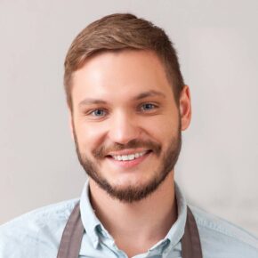 A young man with short brown hair and a neatly trimmed beard smiles warmly at the camera. He is wearing a light blue shirt and a gray apron against a simple, neutral background.