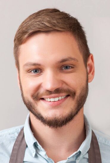 A young man with short brown hair and a neatly trimmed beard smiles warmly at the camera. He is wearing a light blue shirt and a gray apron against a simple, neutral background.