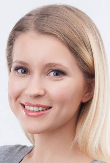 A young woman with long blonde hair smiles at the camera. She is wearing light makeup and a light-colored top. The background is plain and white.