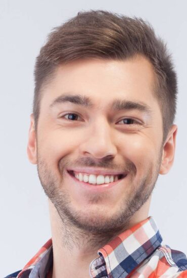 A smiling young man with short brown hair and a beard, wearing a checkered shirt in shades of blue, red, and white, is looking directly at the camera against a light gray background.